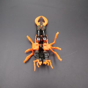 May include: An orange and black toy insect with multiple legs and a large pincer-like head. The toy is made of plastic and has a detailed design, resembling a beetle. The toy is on a gray surface.