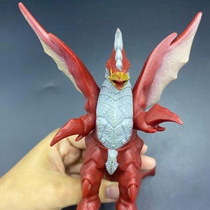 May include: A red and gray toy monster with large wings, a spiked head, and a textured chest. The toy has a bird-like appearance with a yellow beak and is held in a person's hand.