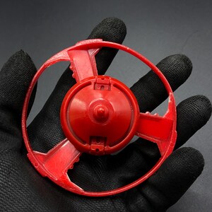 May include: Red plastic toy with a circular base and a ring around the outside. The toy has a central hub with a small, raised point in the center.