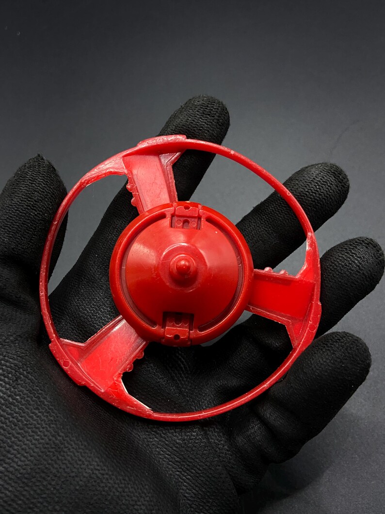 May include: Red plastic toy with a circular base and a ring around the outside. The toy has a central hub with a small, raised point in the center.
