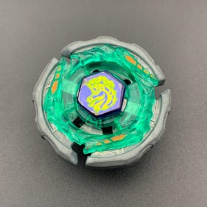 Puede incluir: Un juguete de peonza circular con un anillo exterior gris metálico y una sección central de plástico verde translúcido. En el centro hay un perno hexagonal morado con una ilustración gráfica amarilla de una cabeza de caballo.