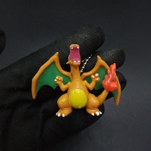May include: A small, orange Charizard figurine with green wings, a yellow belly, and a red flame on its tail. The figurine has white accents on its feet and a silver chain for hanging.