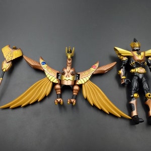 May include: A detailed action figure set featuring a black and gold armored character, a golden bird-like creature with outstretched wings, and a staff. The figures are set against a dark gray background, showcasing intricate design and detail.