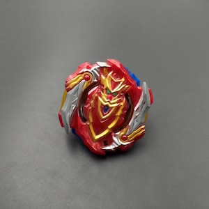 Puede incluir: Un trompo rojo, dorado, plateado y azul. El juguete tiene un diseño complejo con múltiples capas y detalles intrincados. El centro presenta un emblema dorado con detalles verdes. Los bordes exteriores son rojos con elementos plateados y dorados.