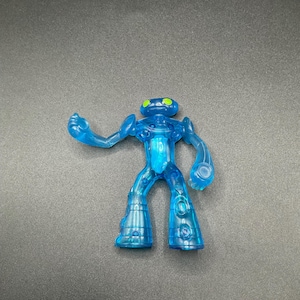 Könnte beinhalten: Eine durchscheinende blaue Actionfigur eines humanoiden Aliens mit grünen Augen. Die Figur hat ein stilisiertes Design mit ausgestellten Beinen und Armen in einer dynamischen Pose. Das Spielzeug besteht aus klarem, glänzendem Kunststoff.