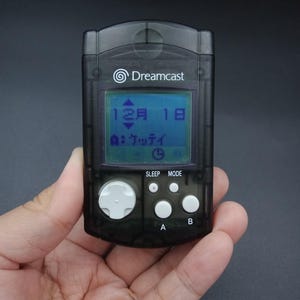 May include: A black Dreamcast Visual Memory Unit (VMU) handheld game device. The screen displays the date "12月 1日". It has a directional pad, A and B buttons, and Sleep/Mode buttons. The device is held in a hand.