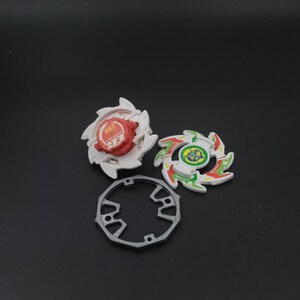 May include: Three colourful spinning tops with a grey ring on a dark background. Two tops are white with red and green accents, and the third is grey. The tops have a circular design with sharp edges.