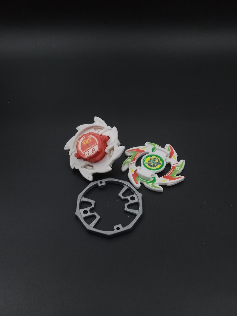 May include: Three colourful spinning tops with a grey ring on a dark background. Two tops are white with red and green accents, and the third is grey. The tops have a circular design with sharp edges.