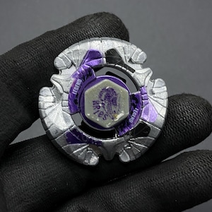 Może przedstawiać: Srebrny i fioletowy Beyblade z czarno-białym wzorem. Beyblade ma fioletowe centrum z białym sześciokątnym elementem przedstawiającym smoka. Beyblade ma napis "Legend" na boku.