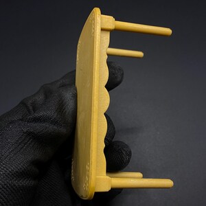 May include: A small, mustard-yellow wooden table with a scalloped edge and three cylindrical legs. The table has decorative floral accents and is designed for a dollhouse or miniature setting.