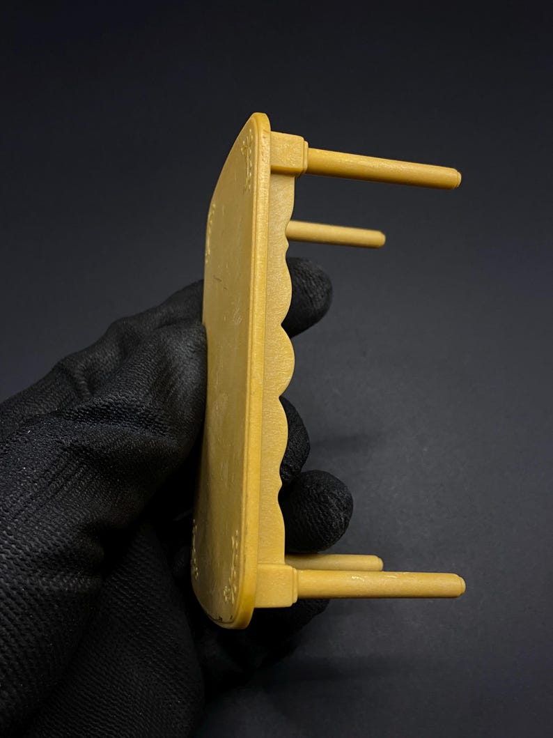 May include: A small, mustard-yellow wooden table with a scalloped edge and three cylindrical legs. The table has decorative floral accents and is designed for a dollhouse or miniature setting.