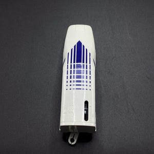 May include: A white and blue air freshener with a geometric design. The air freshener has a rectangular shape with a rounded top and a small opening on the side. The design features vertical blue lines and a blue triangle.