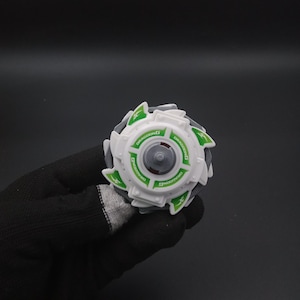May include: A white and grey toy with green detailing and the word "DRAGOONG" printed on it. The toy has a circular design with a central grey component and is held in a black gloved hand.