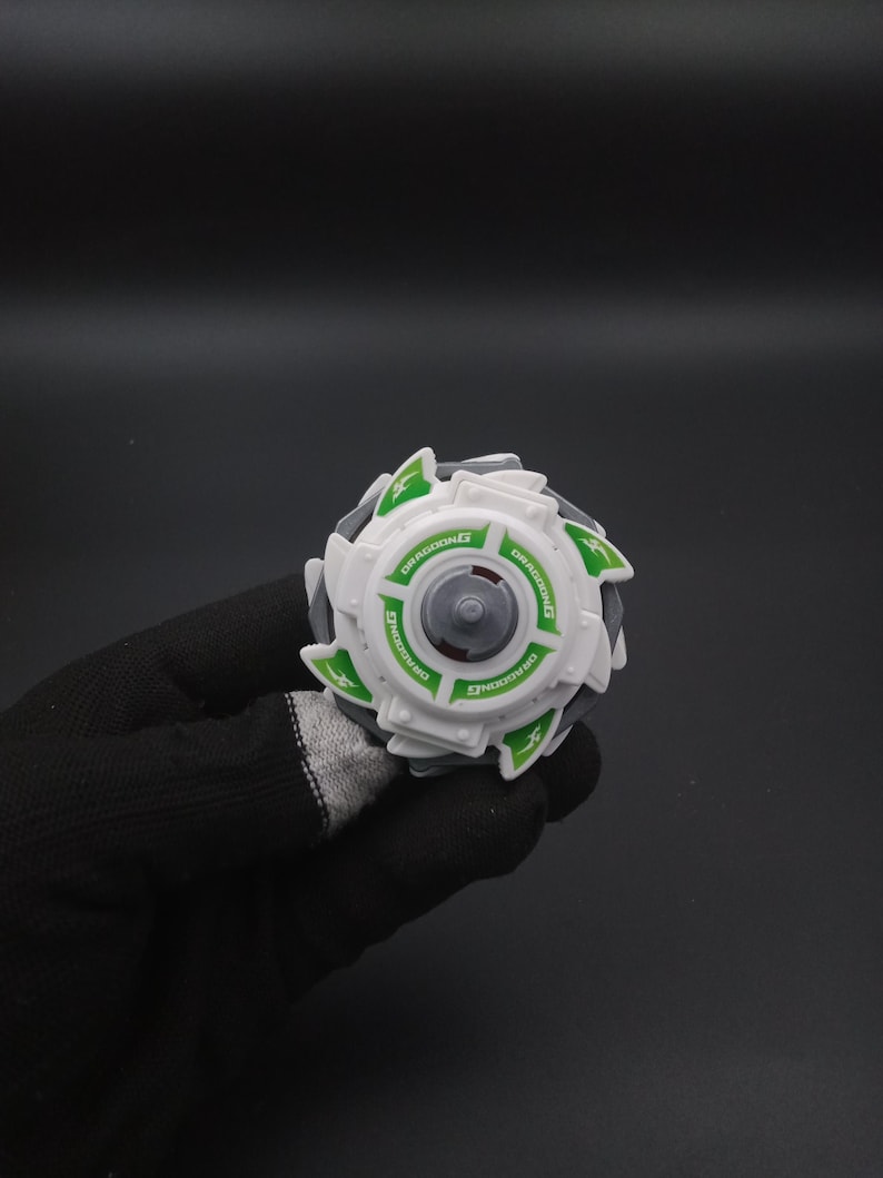 May include: A white and grey toy with green detailing and the word "DRAGOONG" printed on it. The toy has a circular design with a central grey component and is held in a black gloved hand.