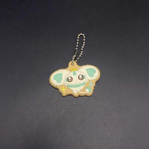 K&ouml;nnte beinhalten: Ein skurriler Schl&uuml;sselanh&auml;nger mit einem Cartoon-Charakter-Design. Der Anh&auml;nger ist wie ein Keks geformt, mit einer hellbraunen Basis, einem niedlichen Gesicht, mintgr&uuml;nen Akzenten und einer kleinen goldenen Krone. Er enth&auml;lt eine silberne Kugelkette zur Befestigung.