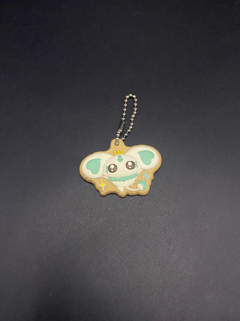 K&ouml;nnte beinhalten: Ein skurriler Schl&uuml;sselanh&auml;nger mit einem Cartoon-Charakter-Design. Der Anh&auml;nger ist wie ein Keks geformt, mit einer hellbraunen Basis, einem niedlichen Gesicht, mintgr&uuml;nen Akzenten und einer kleinen goldenen Krone. Er enth&auml;lt eine silberne Kugelkette zur Befestigung.