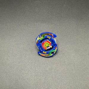May include: A blue and yellow spinning top toy with the word "Dranzer" printed on it. The toy has a circular design with multiple layers and colourful accents. It is a collectible item.