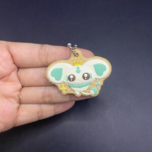 K&ouml;nnte beinhalten: Ein Schl&uuml;sselanh&auml;nger mit einem Cartoon-Charakter mit wei&szlig;em Gesicht, mintgr&uuml;nen Akzenten und einer goldenen Krone. Der Charakter hat gro&szlig;e Augen und befindet sich vor einem hellbraunen Hintergrund. Der Schl&uuml;sselanh&auml;nger ist an einer silbernen Kette befestigt.