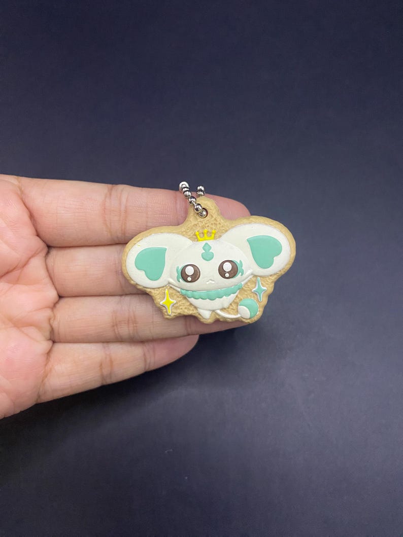 K&ouml;nnte beinhalten: Ein Schl&uuml;sselanh&auml;nger mit einem Cartoon-Charakter mit wei&szlig;em Gesicht, mintgr&uuml;nen Akzenten und einer goldenen Krone. Der Charakter hat gro&szlig;e Augen und befindet sich vor einem hellbraunen Hintergrund. Der Schl&uuml;sselanh&auml;nger ist an einer silbernen Kette befestigt.