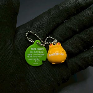 May include: A yellow frog keychain with red eyes and a green tag. The tag reads "HOT FROG" and includes Japanese text. The keychain is attached to a silver chain. The frog has the Japanese text "あったか〜い" on its belly.
