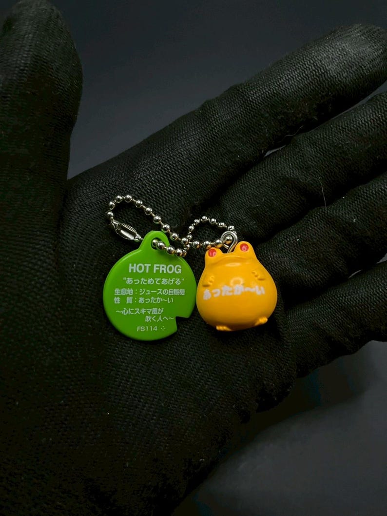 May include: A yellow frog keychain with red eyes and a green tag. The tag reads "HOT FROG" and includes Japanese text. The keychain is attached to a silver chain. The frog has the Japanese text "あったか〜い" on its belly.