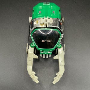 Könnte beinhalten: Grünes und silbernes Spielzeugfahrzeug mit einer krallenartigen Front. Das Spielzeug hat einen grünen Oberteil, einen silbernen Mittelteil und cremefarbene Krallenarme. Das Fahrzeug hat schwarze Ketten und kleine Räder. Das Spielzeug steht auf einer schwarzen Oberfläche.