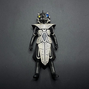 May include: Black and white action figure with a geometric design. The figure has a black helmet with a blue and yellow design. The figure is standing on a gray background.
