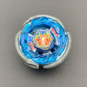 Könnte beinhalten: Ein kreisförmiges Kreiselspielzeug mit einem äußeren Ring aus silberfarbenem Metall und einer inneren Schicht aus durchscheinendem blauem Kunststoff. Die Mitte zeigt ein sechseckiges Emblem mit einem roten und gelben Pferdemotiv und dem roten Schriftzug Pegasus.