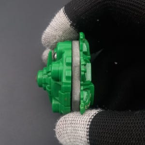May include: A green and gray toy, possibly a spinning top or similar game piece. The toy is held by a gloved hand, showing its side profile. The green plastic has a detailed, geometric design.