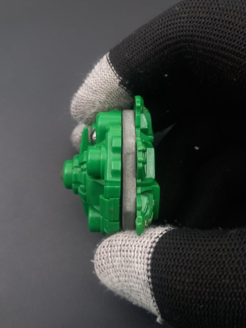 May include: A green and gray toy, possibly a spinning top or similar game piece. The toy is held by a gloved hand, showing its side profile. The green plastic has a detailed, geometric design.