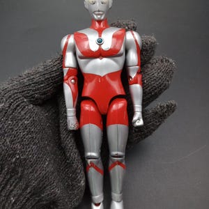 May include: A vintage action figure of a superhero with a silver head and body, red torso and legs. The figure has a blue gem on its chest and is posed upright. The figure is a collectible toy.