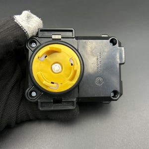 May include: A black plastic toy component with a yellow circular centre. The component has a rectangular shape with rounded edges and several screw holes. The text "OTIS OTOMY MADE IN VIETNAM" is visible on the side.