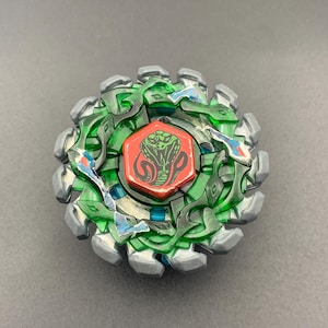 Puede incluir: Un juguete de peonza circular con un anillo exterior de metal plateado de bordes dentados y un marco de plástico verde translúcido. En el centro, un perno hexagonal rojo muestra una ilustración gráfica negra de una cabeza de cobra.
