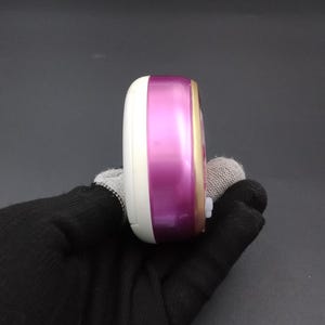 Puede incluir: Dispositivo electr&oacute;nico port&aacute;til blanco, morado y dorado. El dispositivo tiene una forma rectangular redondeada y un peque&ntilde;o bot&oacute;n blanco en el lateral. Se sostiene en una mano enguantada de negro. Probablemente sea un peque&ntilde;o gadget electr&oacute;nico.