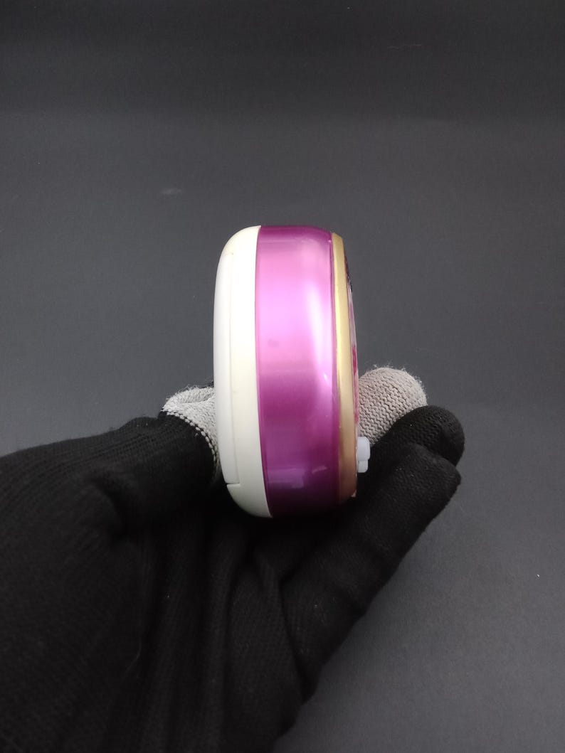 Puede incluir: Dispositivo electr&oacute;nico port&aacute;til blanco, morado y dorado. El dispositivo tiene una forma rectangular redondeada y un peque&ntilde;o bot&oacute;n blanco en el lateral. Se sostiene en una mano enguantada de negro. Probablemente sea un peque&ntilde;o gadget electr&oacute;nico.