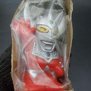 Può includere: Action figure rossa e argentata di un personaggio con un volto sorridente, confezionata in un sacchetto di plastica trasparente e in una scatola di cartone. La figura ha un corpo rosso, testa argentata e dettagli dorati. La figura è in una scatola di cartone.