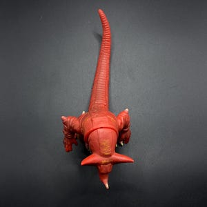 May include: Red plastic toy dinosaur with a long tail and a spiky head. The dinosaur is posed in a standing position with its head lowered.