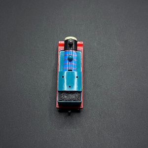 May include: A toy train with a red chassis, teal body, and black smokestack. The train has blue and red stripes on the top and a small white dome. The toy is on a dark gray surface.