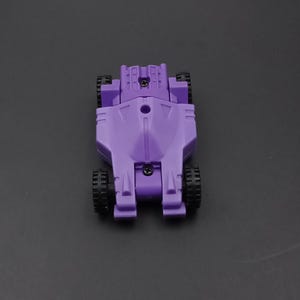 May include: A purple toy car with black wheels. The toy car has a streamlined design with a central body and four wheels. The wheels are black and the body is a solid purple color. The toy is viewed from above.