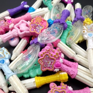 May include: A collection of colorful plastic wands with star-shaped designs and glitter accents. The wands have white handles and clear plastic wings.
