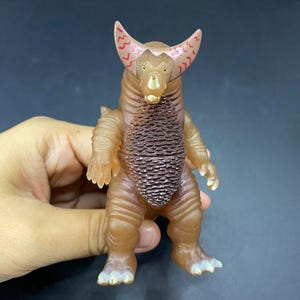 May include: A brown action figure of a monster with large horns and a textured chest. The figure has a light brown body with darker brown accents and red patterns on the horns. The figure is held by a hand.