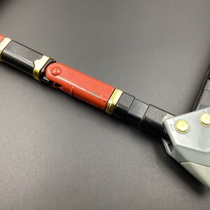 May include: A red, black, and gold toy weapon with a gray plastic tip. The weapon is made of multiple sections that can be connected together.