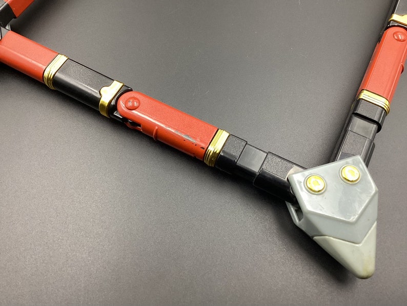 May include: A red, black, and gold toy weapon with a gray plastic tip. The weapon is made of multiple sections that can be connected together.