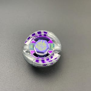 Puede incluir: Un juguete de peonza circular con un anillo exterior de metal plateado y detalles en morado. El centro incluye un perno hexagonal morado con un gráfico verde claro de una criatura estilizada y texto. El diseño tiene un aspecto mecánico y en capas.
