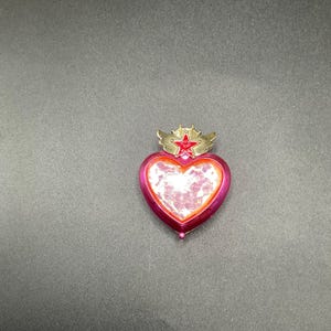 May include: A heart-shaped, pink and red decorative object with a star and crown design. The object has a gold-colored crown and star on top. The heart has a clear, textured center and a red border. The object is on a dark gray surface.