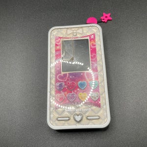Puede incluir: Un juguete Smart Pod Touch plateado y rosa con una pantalla agrietada. El juguete tiene una estrella rosa y una perla rosa adjunta en la parte superior. La pantalla muestra varios iconos y el texto "Smart pod touch".