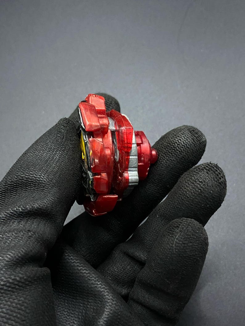 K&ouml;nnte beinhalten: Rotes und silbernes Kreiselspielzeug mit einer durchsichtigen Plastikspitze. Der Kreisel befindet sich in einer Hand, aber die Hand ist nicht sichtbar.