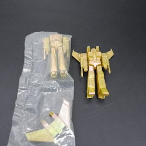 May include: Two gold-colored toy robots, one assembled and one in a plastic bag. The assembled robot has wings and a detailed design. The other robot is unassembled. The toys are on a dark surface.
