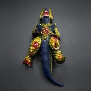 Pode incluir: Um brinquedo de monstro azul e dourado com olhos vermelhos e uma boca bem aberta. O monstro está em pé sobre suas patas traseiras com as patas dianteiras levantadas. O brinquedo é feito de plástico e tem um design detalhado.