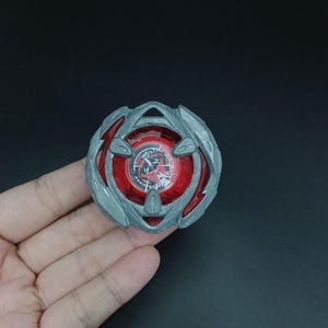 May include: A red and gray spinning top toy with a circular design. The top has a red center with a graphic and is surrounded by a gray, multi-layered outer ring. The toy is held in a hand against a dark background.